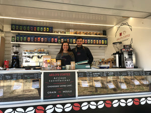 Laura et Arnaud, les gérants et créateurs d'Arlo's Coffee se tiennent debout dans leur coffee truck, présent sur un marché. Au premier plan se trouve les différents cafés.  