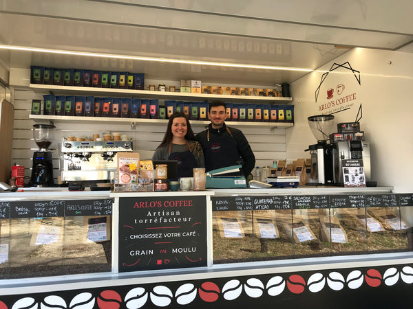 Laura et Arnaud, les gérants et créateurs d'Arlo's Coffee se tiennent debout dans leur coffee truck, présent sur un marché. Au premier plan se trouve les différents cafés.  